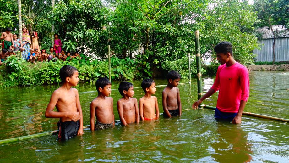 Five young boys at a water safety lesson being taught whilst their families watch from the side of the water